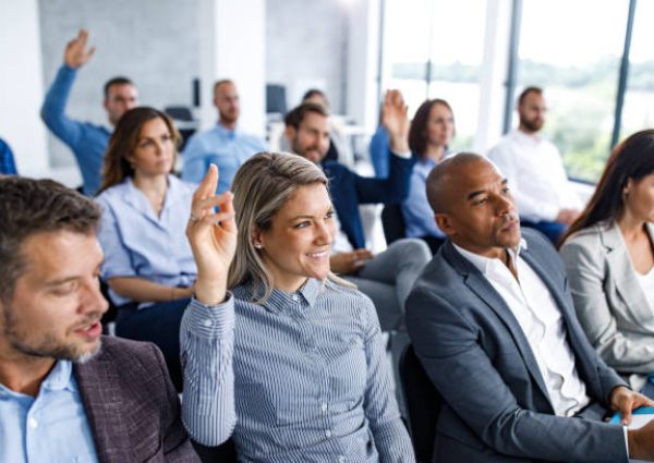 Large group of business people attending an education event in a board room. Focus is on happy woman raising her hand to ask a question.